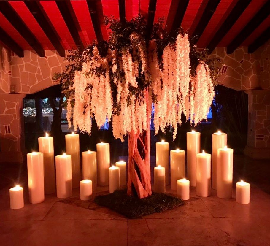 Elegant tree with hanging white flowers, surrounded by tall lit candles in a dimly lit room.